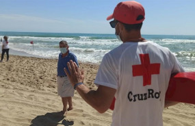 ep un senor pasea por la playa con mascarilla y atiende a lo que le dice un socorrista de la cruz