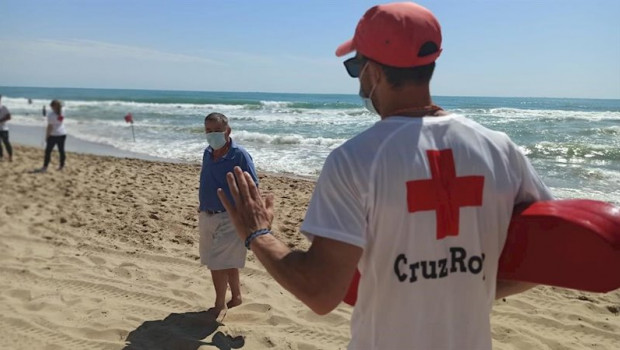 ep un senor pasea por la playa con mascarilla y atiende a lo que le dice un socorrista de la cruz